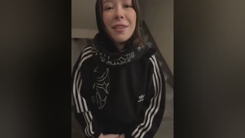 Media: Video of a smiling, fair-skinned woman with brown hair, wearing a black Adidas hoodie with white stripes and a graphic print. She stands in a dimly lit room with neutral walls.