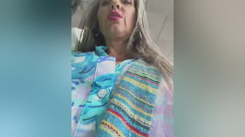 Media: Video of a Caucasian woman with long, wavy, light brown hair, wearing a colorful, striped, knit sweater and a turquoise jacket, in a neutral indoor setting.