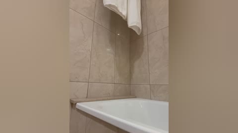 Media: A video of a modern bathroom corner featuring a white bathtub with beige marble tiles, a white towel hanging from a shelf above, and a beige wall.