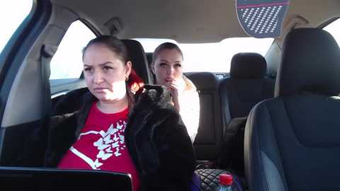 Media: Video of two women in a car: one with dark hair in a ponytail, wearing a red graphic tee, looks bored; the other, with blonde hair, contemplative, in the back seat.