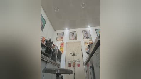 Video of a small, modern kitchen with white walls, recessed ceiling lights, and framed artwork. Stainless steel appliances, a sink, and a small stool are visible. The image is taken from a low angle, creating a perspective effect.