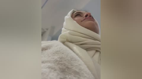 Media: A low-angle video of a person wrapped in a white, fluffy towel, viewed from the neck up, looking upward with a serene expression. The background is blurred, suggesting an indoor setting.