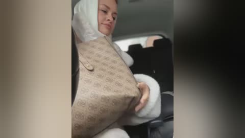 Media: A video captures a woman in a beige, patterned coat and white headscarf, seated in a car, appearing relaxed with a slight smile. The background shows a blurred interior with black seats.