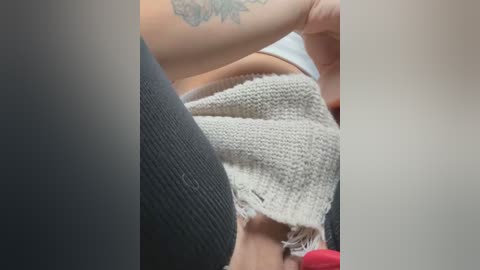 Media: Video of a person wearing a grey, ribbed sweater with frayed edges, partially covering a tattoo on their upper arm. The background is blurred, focusing on the sweater's texture and the person's arm.