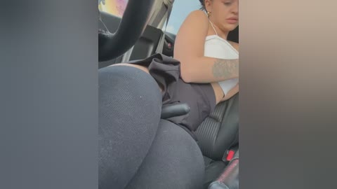 Media: A video of a tattooed woman in a tight black dress, reclining in a car seat, wearing dark leggings. Her right arm rests on the car door, and she appears relaxed.