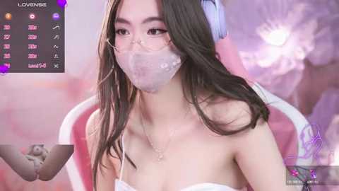 Media: Video of a young Asian woman with long black hair, wearing a clear face mask, white strapless top, and headphones, seated in a pink chair, surrounded by a pastel pink and purple background.