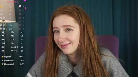 Video of a young woman with long, reddish-brown hair and fair skin, smiling, wearing a grey sweater. Background features dark curtains and a purple chair.