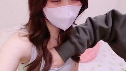 Video of a young Asian woman with long brown hair, wearing a white face mask and gray hoodie, lying on a bed with a white sheet and a pink pillow, showcasing a casual, intimate setting.