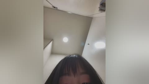 Media: A video of a young woman with long, straight black hair, captured from a low angle, looking up at a ceiling with fluorescent lights and light fixtures.