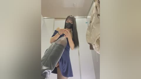 Media: Video of an Asian woman with long black hair and a black mask, wearing a blue off-shoulder dress, standing in a white-walled, dimly lit room, holding a high-heeled shoe in front of her.