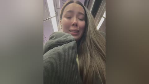 Media: A video of a smiling young woman with long, straight, light brown hair, wearing a grey ribbed sweater, standing in a dimly lit, narrow hallway with fluorescent lighting and white walls.