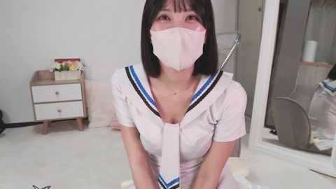 Video of an East Asian woman with a short black bob haircut, wearing a white sailor outfit, face mask, and glasses, kneeling on a white carpet in a modern, minimalist bedroom.