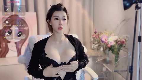 Media: Video of an East Asian woman with fair skin and medium-length black hair, wearing a black bra and unbuttoned jacket, standing in a well-lit room with a camera on tripod, flowers, and an abstract painting in the background.