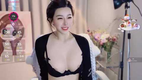 Media: A video of an Asian woman with fair skin and dark hair, wearing a black lace bra, smiling, standing in a room with a TV screen displaying her stats.