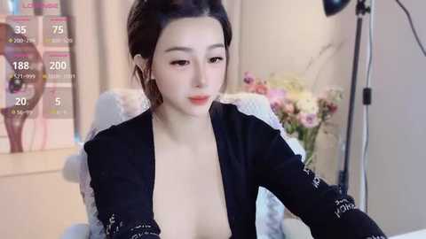 Media: A video of an East Asian woman with fair skin and dark hair, wearing a black blouse, sitting in a chair with a pink vase and floral backdrop.