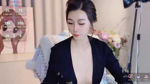 Media: Video of an East Asian woman with pale skin, black hair in a bun, wearing a low-cut black top, sitting in a soft-lit room with a floral arrangement and a vintage chair in the background.