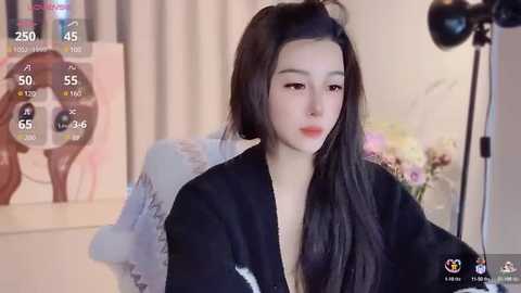 Media: A video of a young East Asian woman with long black hair, wearing a black blouse, sitting in a dimly lit room with a flower arrangement in the background.