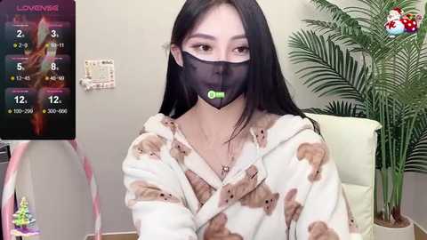 Video of an Asian woman with long black hair, wearing a black face mask, white pajama top with teddy bear print, and a potted plant in the background.