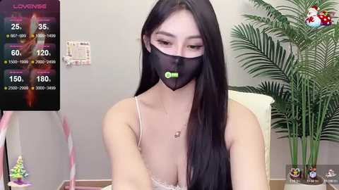 A young Asian woman with long black hair, wearing a black face mask, white lace lingerie, and a green necklace, sits indoors near a green plant, with a live-streaming overlay displaying stats and viewers.