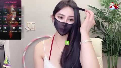 Media: Video of an East Asian woman with long black hair, wearing a white lace bralette, black face mask, and green ID card, adjusting a pink ribbon. Background shows a plant, digital thermometer, and a TV displaying a weather forecast.