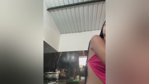 Media: Video of a woman in a pink tank top, standing in a small, dimly-lit kitchen with black tiled walls and a metallic ceiling.