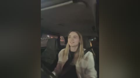 Video of a smiling young woman with long blonde hair, wearing a black top and white jacket, driving a car at night.