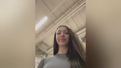 A video of a young woman with long brown hair, wearing a grey top, standing in a dimly lit, industrial room with exposed pipes and a ceiling grid.