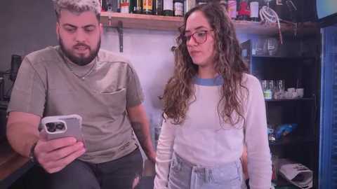 Media: Video of a bearded man in a gray t-shirt, and a curly-haired woman in glasses, holding a phone, in a cluttered, dimly lit bar.