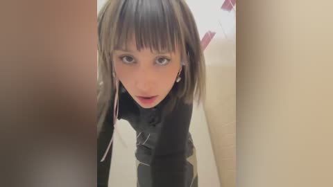 Media: A video of a young Asian woman with straight, dark hair and light skin, wearing a black leather jacket, leaning forward in a dimly lit, narrow hallway with beige walls and a tiled floor.