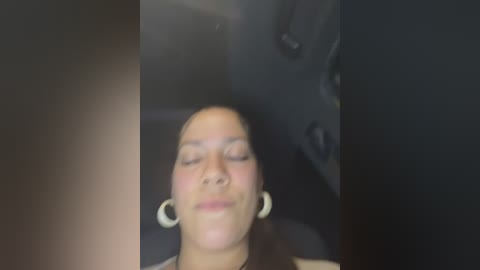 Media: Video of a serene, light-skinned woman with a bald head, wearing large hoop earrings, lying in a car seat, eyes closed, and looking relaxed.