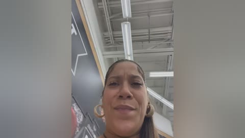 Media: A video of a young Black woman with light brown skin and long, straightened hair. She wears large hoop earrings, a light-colored top, and stands in an industrial room with white walls and exposed beams.