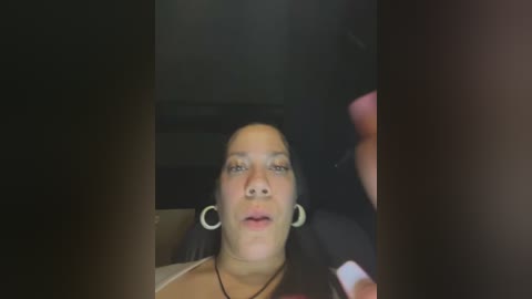 Media: A video of a woman with medium skin tone, long dark hair, hoop earrings, and a neutral expression, sitting in a dark car.