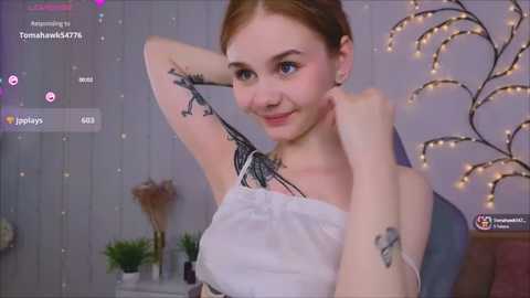 Media: Video of a young woman with fair skin, brown hair, and tattoos on her arm, wearing a white dress, smiling, in a cozy, softly lit room with fairy lights and plants.