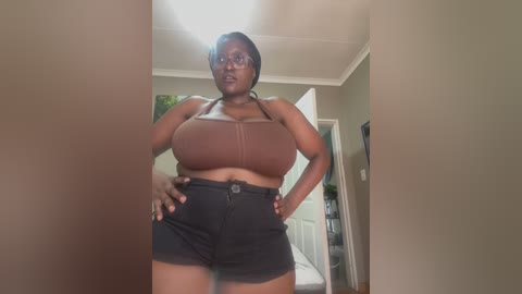 Media: A video of a confident Black woman with a medium complexion, wearing a brown sports bra and high-waisted black shorts, standing in a modern, minimalist home with beige walls and a white door.