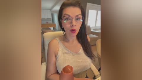 A video of a fair-skinned woman with long brown hair, wearing glasses, and a white ribbed tank top, holding an erect penis. She sits in a modern, beige-colored living room.