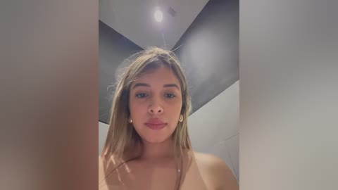 Media: Video of a young, light-skinned woman with straight, light brown hair, wearing a topless outfit. She has a neutral expression and is standing in a modern, minimalist room with gray walls and a recessed ceiling light.