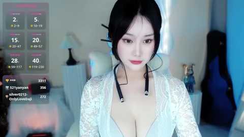 Media: Video of a young Asian woman with fair skin, dark hair styled in a bun, wearing a revealing white lace top that exposes cleavage. Background shows a bedroom with a bed, lamp, and guitar.