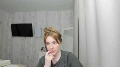 Media: Video of a fair-skinned, red-haired woman with a neutral expression, applying makeup in a minimalist, white-tiled bathroom with a flat-screen TV and white curtains.