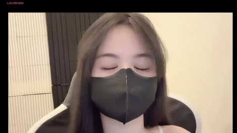 Media: Video of an Asian woman with long, straight, dark hair, wearing a black face mask and a white top, sitting in a modern chair. Background features a black and white folding screen and a beige wall.