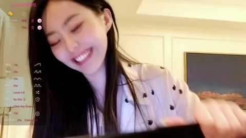 Media: Video of a smiling East Asian woman with long black hair, wearing a white blouse, in a brightly lit room with a large window and wall clock.