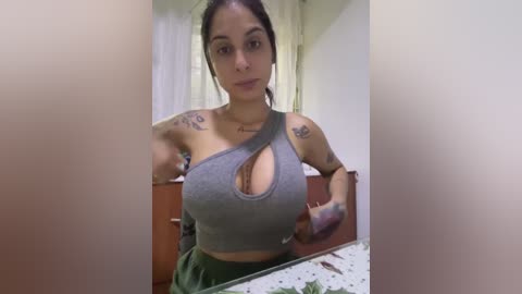 Video of a young woman with light brown skin and medium-length dark hair, wearing a gray crop top revealing cleavage, tattoos on arms, standing in a kitchen.