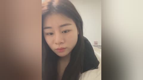 Media: A video of an Asian woman with long black hair and fair skin, wearing a black shirt, looking pensive in a dimly lit room with white walls.