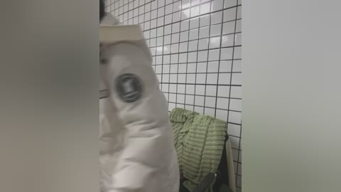 Media: Video of a white, quilted jacket partially obscured by a fogged-up glass door, set against a tiled bathroom wall with a green, textured towel hanging on a hook.