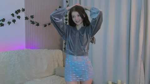 Video of a young woman with long brown hair, wearing a shiny, blue sequin skirt and a gray hoodie, standing in a modern, dimly-lit living room with white walls and green leaf decals.