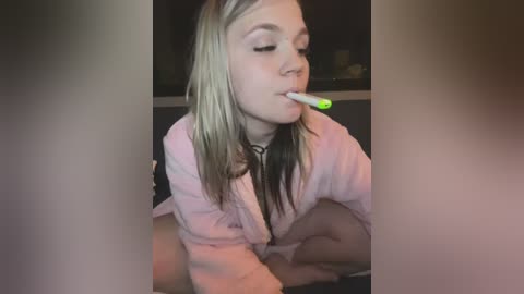 Media: Video of a blonde young woman in a pink robe, with a green toothbrush in her mouth, leaning against a dark surface, blurry background.