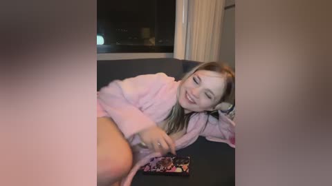 Media: A video of a young woman with long blonde hair, wearing a pink robe, lying on a dark surface, smiling while holding a phone.