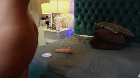 Video of a modern bedroom with a teal, tufted headboard, a gray bed, two pillows, a beige nightstand, a lamp, and a dildo on the bed.