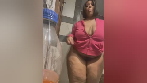 Media: Video of a plus-sized woman with short black hair, wearing a pink zip-up hoodie, standing in a bathroom with a clear plastic bottle and a pink wall in the background.