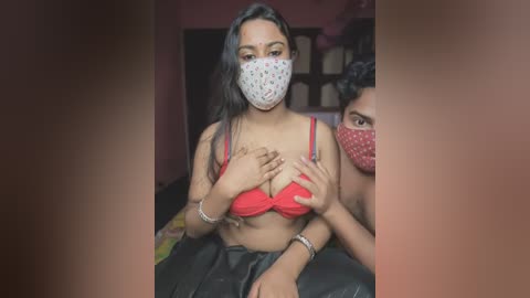 Media: Video of a South Asian woman with medium skin tone and long black hair, wearing a polka-dotted face mask and red bra, holding her breasts, with another person partially visible behind her.