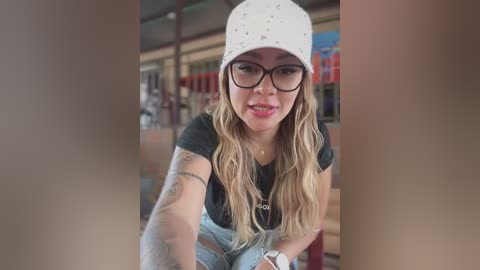 Media: Video of a smiling woman with long, wavy blonde hair, wearing glasses, a white bucket hat, black t-shirt, and ripped jeans, seated in a library, surrounded by bookshelves.